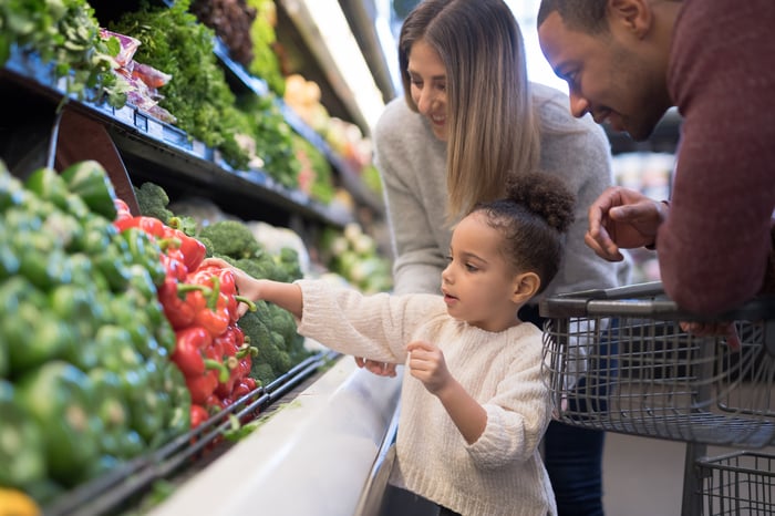 A child reaching for a bell pepper in a grocery store, with their parents watching. 